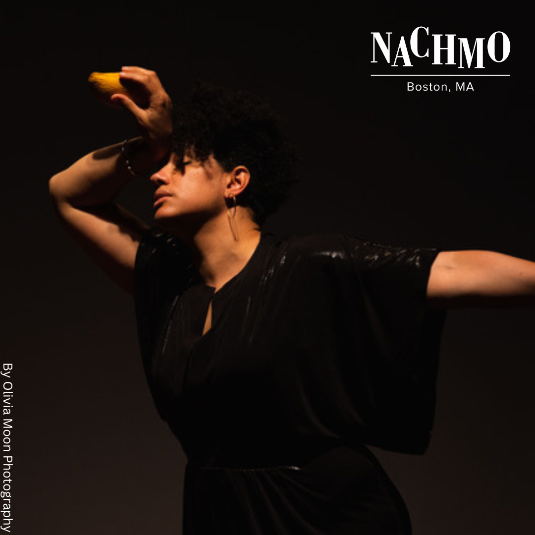 Jess laments in profile, dancing in her solo, Surrender. She perches the back of her wrist over her head while holding a mango, and extends her other arm to the side. She wears a black shiny wide sleeved jumpsuit, gold hoop earrings and silver bangles. 