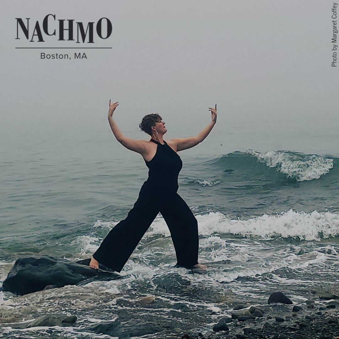 White woman in a black jumpsuit posing on a rock in the ocean with arms open over her head